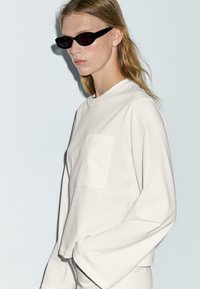Witte lange mouwen top met een ronde hals en een voorzak; gecombineerd met elegante zwarte zonnebril; het haar van het model is golvend en lichtgekleurd.