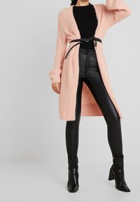 Lång rosa stickad cardigan med öppen front, texturiserad tyg och ribbad ärm, parat med svarta högmidjade läderleggings och ankelstövlar.