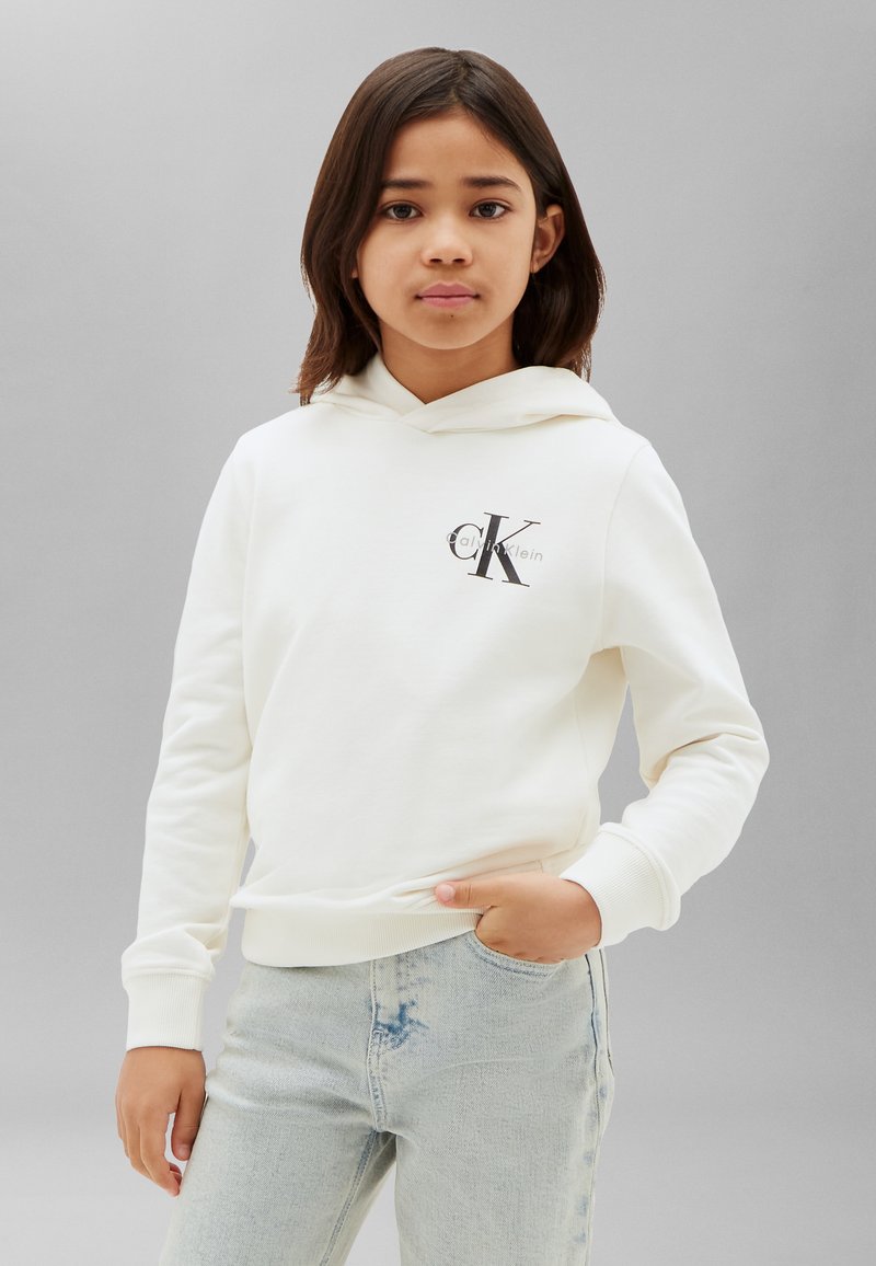 Κορίτσι που φοράει λευκό hoodie Calvin Klein και ανοιχτόχρωμο τζιν, στέκεται με το ένα χέρι στην τσέπη μπροστά σε γκρι απλό φόντο.