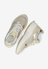 Beige Sport Schuhe mit einem Obermaterial aus Mesh, metallischen Akzenten und weißen Schnürsenkeln. Sie verfügen über eine dicke, weiße Sohle und strukturierte Details an den Seiten.