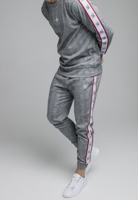 Grau gemusterter Trainingsanzug mit einem Sweatshirt im Crew-Neck-Stil und tapered Hosen mit roten und weißen Seitenstreifen. Kombiniert mit weißen Sneakers.
