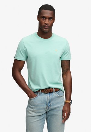 Mann trägt hellgrünes kurzärmeliges T-Shirt, hellblaue Jeans, braunen geflochtenen Gürtel und Armbanduhr, steht mit einer Hand in der Tasche.