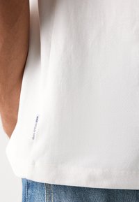 Fehér pamut póló sima textúrával, az alján egy kis kék címkével, amelyen az áll: "Marc O'Polo DENIM". Lazán illeszkedő fazon.