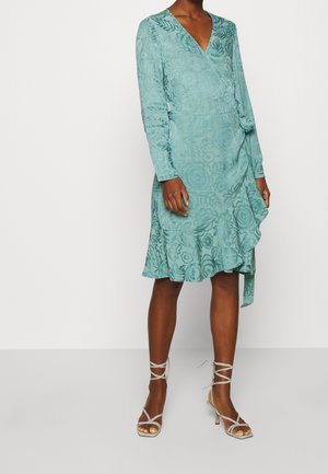 Femme portant une robe portefeuille florale bleu sarcelle à manches longues, associée à des sandales blanches à talons hauts à lanières, debout devant un fond uni.