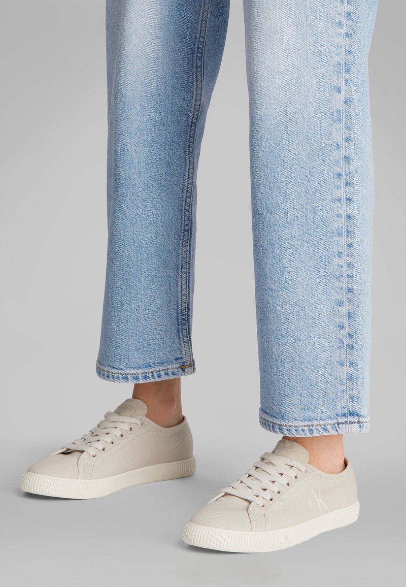 Persoon staand, gekleed in lichtblauwe spijkerbroek en beige canvas sneakers met witte rubberen zolen, tegen een grijze achtergrond.