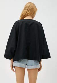 Schwarzes, locker sitzendes Oberteil mit weiten Ärmeln, aus leichtem Material, mit abgerundetem Saum und einer sauberen Rückennaht. Kombiniert mit Jean-Shorts.