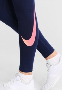 Person klädd i marinblå leggings med en stor rosa swoosh och vita sneakers med vita strumpor, stående mot en enkel ljus bakgrund.