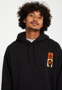 Zwarte hoodie met een vooraan afbeelding met oranje, gele en zwarte elementen. Gemaakt van zachte stof, heeft het een verstelbare trekkoordkap.
