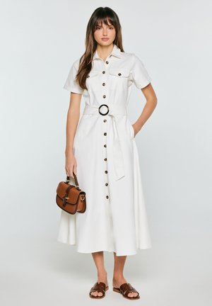Kobieta ubrana w białą midi sukienkę na guziki z paskiem, trzyma brązową torebkę i nosi brązowe sandały wsuwane, stoi na jednolitym tle.