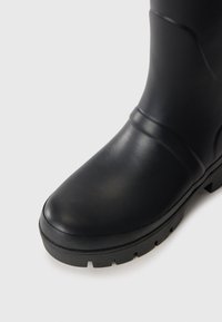 Close-up of a black rubber boot with a rounded toe and thick, ridged sole on a white background.