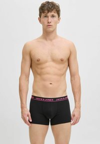 Hemdloser Mann mit heller Haut und lockigem Haar, der schwarze Boxershorts mit einem pinken "JACK & JONES"-Bund trägt und vor einem einfarbigen Hintergrund steht.