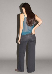 Osoba má na sebe priesvitný teal top bez rukávov, ktorý je kombinovaný s voľnými tmavosivými džínsami s dvoma zadnými vreckami. Pozadie je sivé.