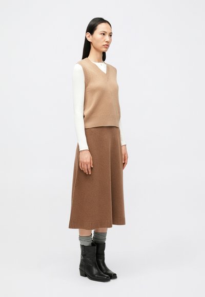 Naine, kes seisab, kandes beežikat varrukateta kampsunivesti, sobivat midi-seelikut, valget pika varrukaga särki, halli sokke ja musti pahkluu-saapaid.