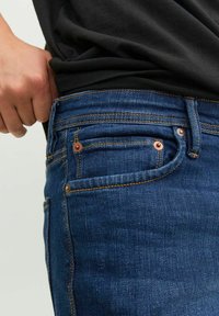Jeans de denim azul oscuro con una textura suave, que presentan remaches de cobre, costuras en contraste y un pequeño bolsillo delantero.