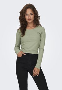 Långärmad, ribbad grön topp med rund halsringning och vågig nederkant, matchad med högmidjade svarta byxor. Strukturen i tyget är synlig.