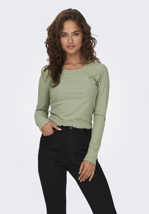 Langmouw, geribbelde groene top met een ronde hals en golvende zoom, gecombineerd met hoge zwarte broek. Gestructureerde stof is zichtbaar.