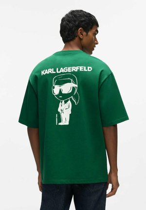Homme portant un t-shirt vert oversize avec un personnage de dessin animé blanc et le texte ""KARL LAGERFELD"" au dos.