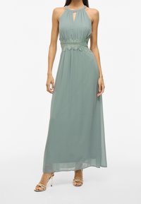 Lång, ärmlös grön klänning med halterneck, med nyckelhålskärning och spetsdetaljer vid midjan, flödande tyg och en ledig passform.