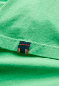 Grünes Baumwoll-T-Shirt mit sichtbarem Markenlabel auf navyblauem Hintergrund und roten, weißen sowie grünen Streifen am Saum.