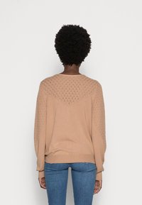 Suéter de punto beige con un patrón texturizado en los hombros y las mangas, con un corte holgado y puños acanalados, combinado con jeans azules.