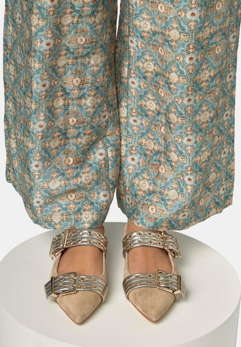 Pieds portant des ballerines beiges à bout pointu avec des brides à double boucle métallique, associées à un pantalon ample à motifs floraux bleus et beiges.