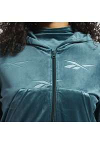 Gros plan d'une personne portant un sweat à capuche zippé bleu sarcelle avec des logos Reebok en relief et de longs cheveux bouclés visibles sur les épaules.