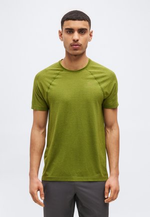 Jeune homme debout, portant un t-shirt moulant à manches courtes texturé de couleur vert olive et un pantalon gris foncé, sur un fond clair uni.