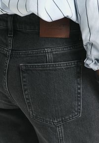 Jean en denim foncé avec une texture lisse, une poche arrière et une étiquette en cuir de la marque GANT à la taille. Présente un design ajusté.