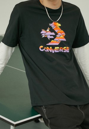 T-shirt en coton noir avec un graphique de nuage coloré et le texte "CONVERSE". Coupe décontractée, manches courtes, superposé avec un sous-vêtement à manches longues blanc en dessous.
