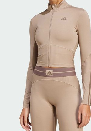 Femme portant un top de sport beige ajusté avec fermeture éclair et un legging taille haute assorti avec logos Adidas sur la poitrine et la ceinture.