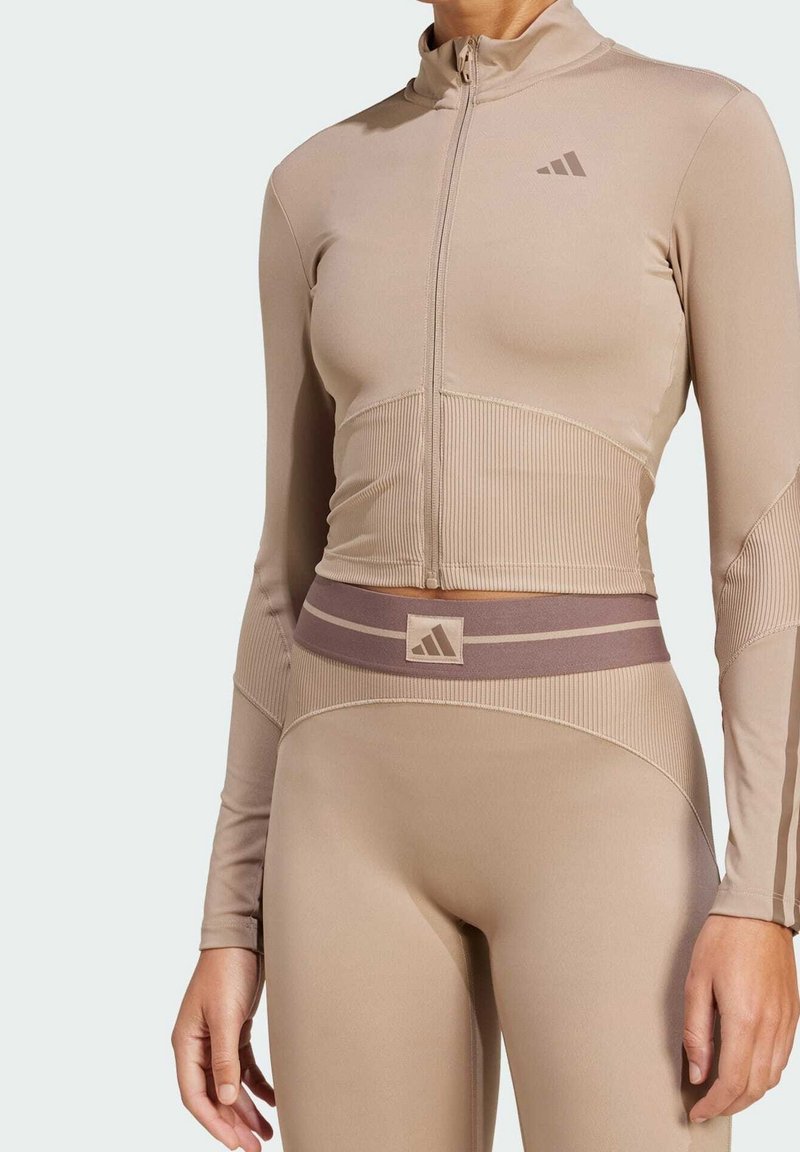 Femme portant un top de sport beige ajusté avec fermeture éclair et un legging taille haute assorti avec logos Adidas sur la poitrine et la ceinture.