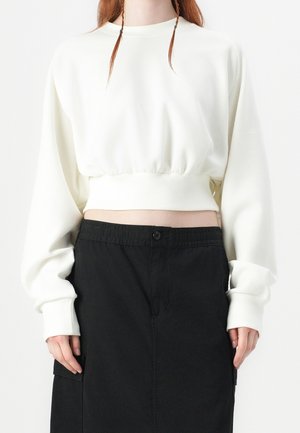 Femme portant un pull court blanc à manches longues et une jupe noire taille haute, montrant le ventre et une partie des cheveux roux.