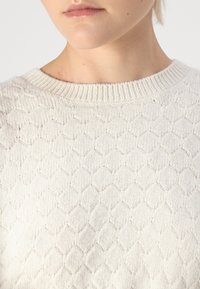 Krema barve pletena majica z teksturiranim satastim vzorcem in rebrastim okroglim izrezom, ki prikazuje mehak in udoben material.