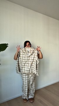 Osoba stojaca v interiéri a drží béžovo-bielu károvanú košeľu s malými motívmi stromov, oblečená v zladených károvaných nohaviciach a hnedých topánkach.