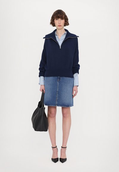 Hanorac navy cu guler cu fermoar, împreună cu o fustă din denim, ținând o geantă neagră tip tote. Prezintă tocuri negre ascuțite și manșete cu dungi albastre.