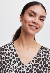Blouse en imprimé léopard avec un décolleté en V, fabriquée en tissu léger. Les couleurs incluent des motifs crème, noir et marron. Texture douce.