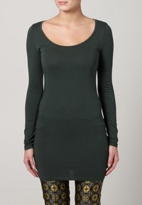 Žena oblečená v přiléhavých tmavě zelených dlouhých šatech a vzorovaných černých a zlatých legínách, stojící proti jednobarevnému světlému pozadí.