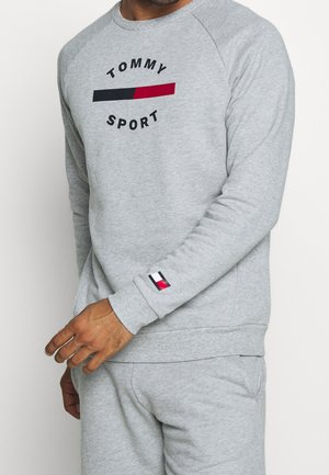 Hombre con sudadera Tommy Sport gris claro y pantalones deportivos a juego, con texto del logo y bloque de color en el pecho y el puño de la manga.