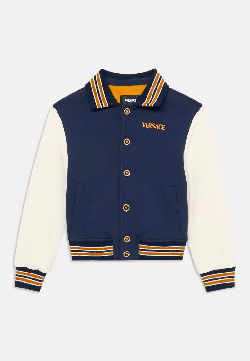Casaco varsity azul-marinho e creme com colarinho e punhos riscadados. Apresenta botões dourados e o logótipo "VERSACE" em laranja. Tecidos suaves, design desportivo.