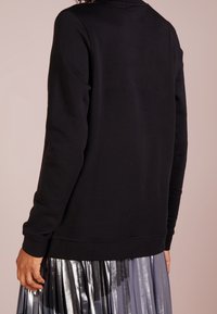 Černá mikina s dlouhými rukávy, kulatým výstřihem a volným střihem, kombinovaná s metalickou plisovanou sukní. Celková matná textura.