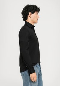 Ung mann med krøllete mørkt hår iført en svart langermet skjorte med knapper og blå jeans, stående i profil mot en ensfarget bakgrunn.