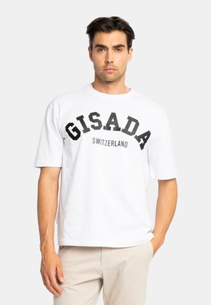 Mann mit dunklem Haar, der ein weißes kurzärmeliges T-Shirt mit dem Text "GISADA SWITZERLAND" und beige Hosen trägt, steht vor einem einfachen Hintergrund.
