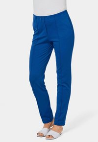 Blauwe gepaste broek van een gladde stof, met voorwervels en zijzakken. Gemaakt met witte sandalen met open tenen.