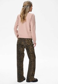 Kobieta z długimi włosami, ubrana w różowy sweter z dzianiny i szerokie spodnie w print w panterkę, stojąca na tle jednolitego białego tła.