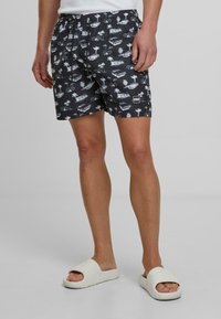 Fekete úszóshort fehér pálmafa és autó mintákkal, rugalmas derékkal, és egy kis logóval a bal oldalon. Fehér strandpapuccsal viselve.