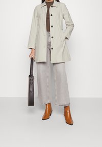 Manteau gris clair longueur genou avec col, boutons sur le devant et poches latérales. Porté sur un haut marron, assorti à un jean clair à jambes larges et des bottines marron.