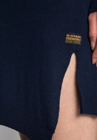 Pull en maille bleu marine avec ourlet côtelé et fente latérale, présentant une étiquette en denim G-Star Originals. Texture lisse avec une coupe décontractée.