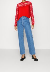 Donna che indossa un top lungo in pizzo rosso brillante, jeans blu a vita alta e décolleté in vernice nera, in piedi contro uno sfondo neutro.