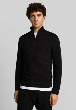 Uomo con capelli ricci che indossa un maglione nero maculato con zip a un quarto sopra una camicia bianca contro uno sfondo grigio semplice.