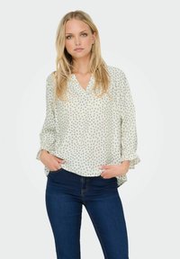 Heledatud blouse, millel on V-kaelus ja kolmeveerandipealne varrukas, millel on tumesinised lillemustrid heledal taustal. Kandmisel tumedate teksadega.